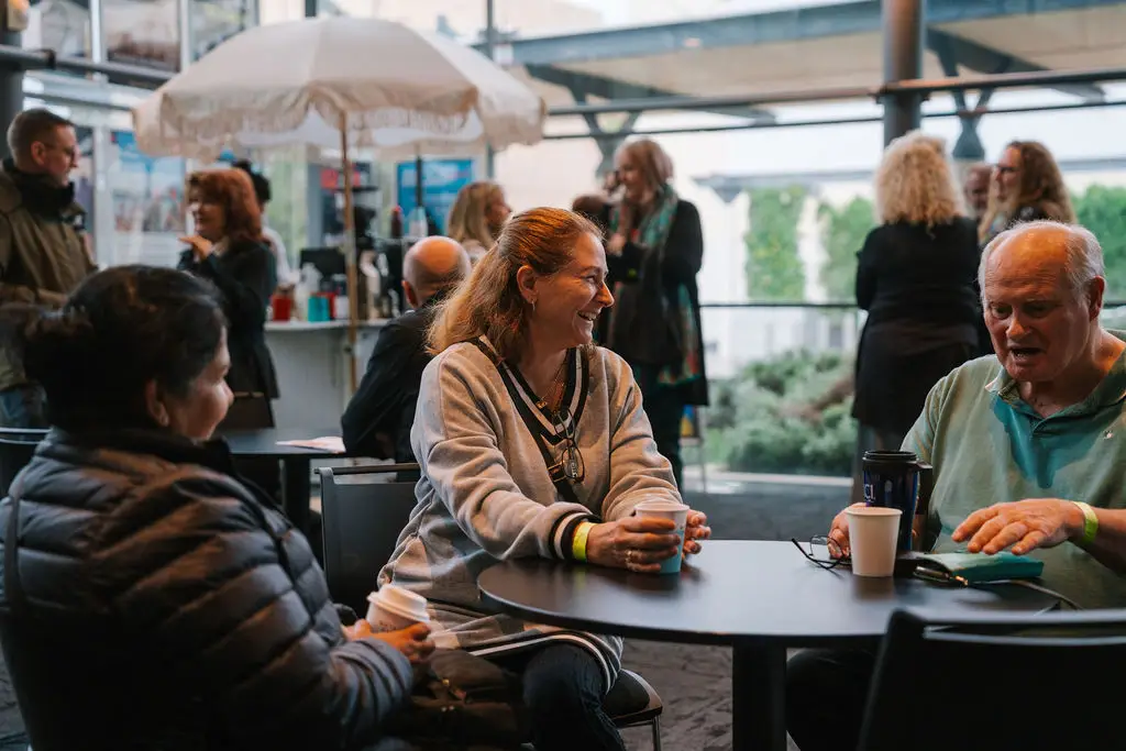 Small group seated at a round table in a lobby area, talking and holding cups.